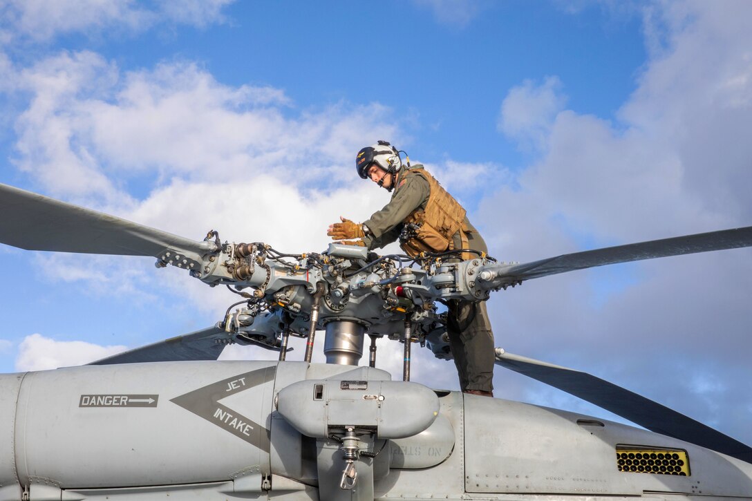 An airman stands on top of a helicopter to perform pre-flight checks