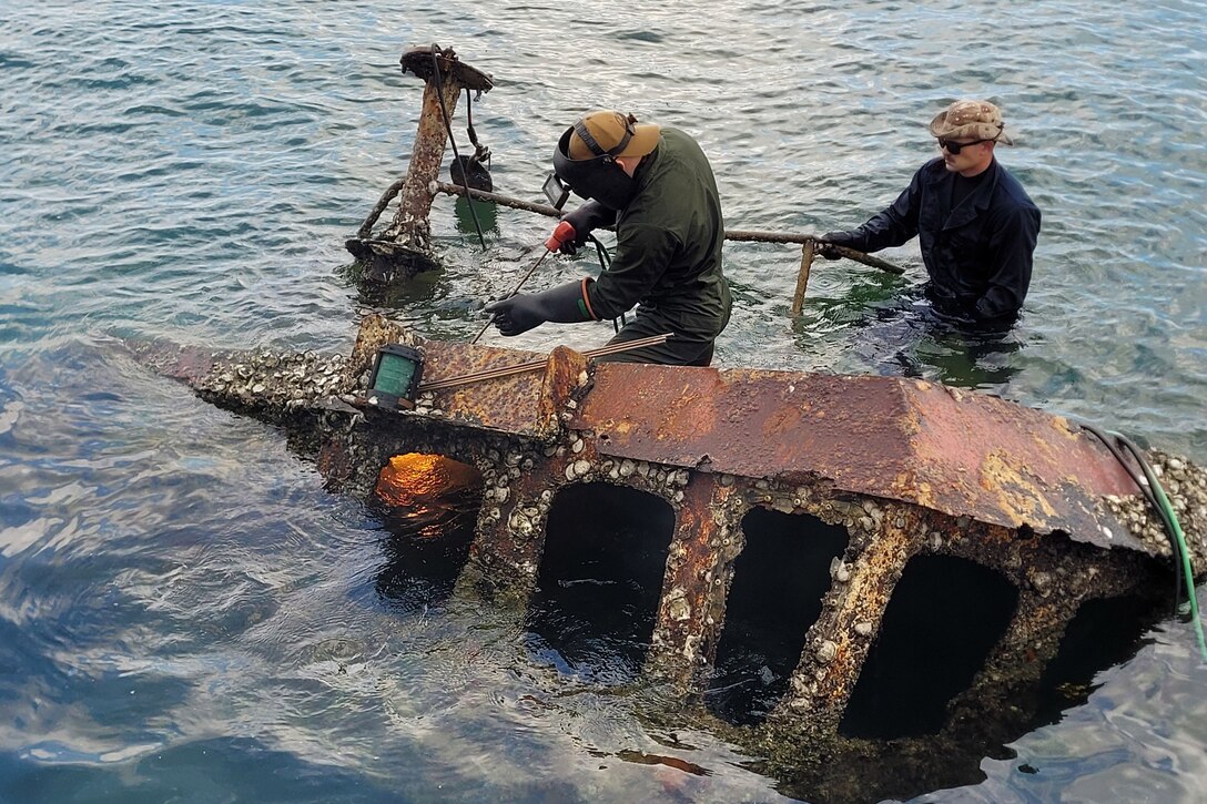 Two sailors use equipment to cut a metal structure in shallow waters.