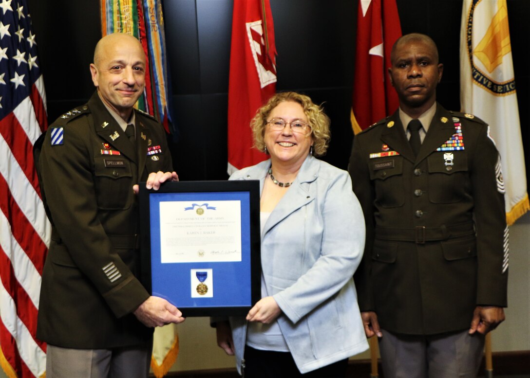 Two men in Army uniforms stand on each side of a woman holding an award being given to her.