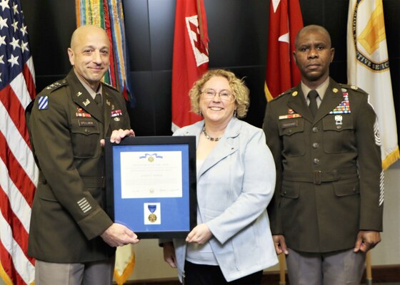 Two men in Army uniforms stand on either side of a woman being handed an award.
