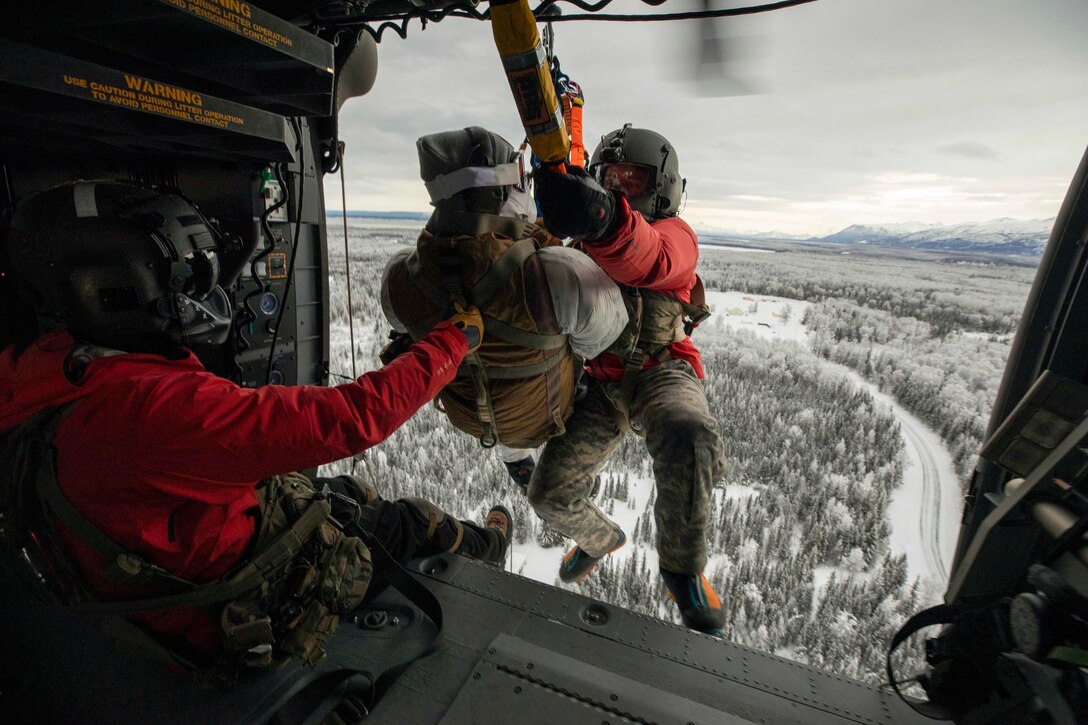 A solider sitting in an airborne helicopter pulls up and a fellow soldier and another person connected by a rope.