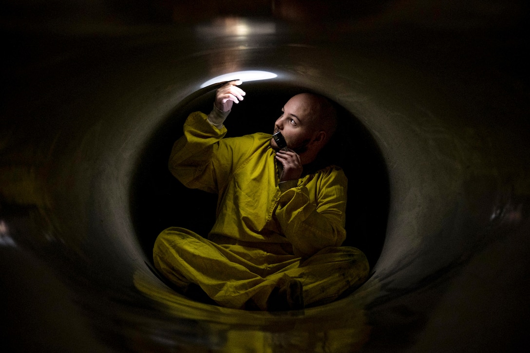 An airman in coveralls holds up a flashlight while sitting inside a round opening in a metal body.