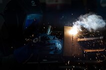 A welder's mask is illuminated by sparks as he practices vertical stick welding in front of him.