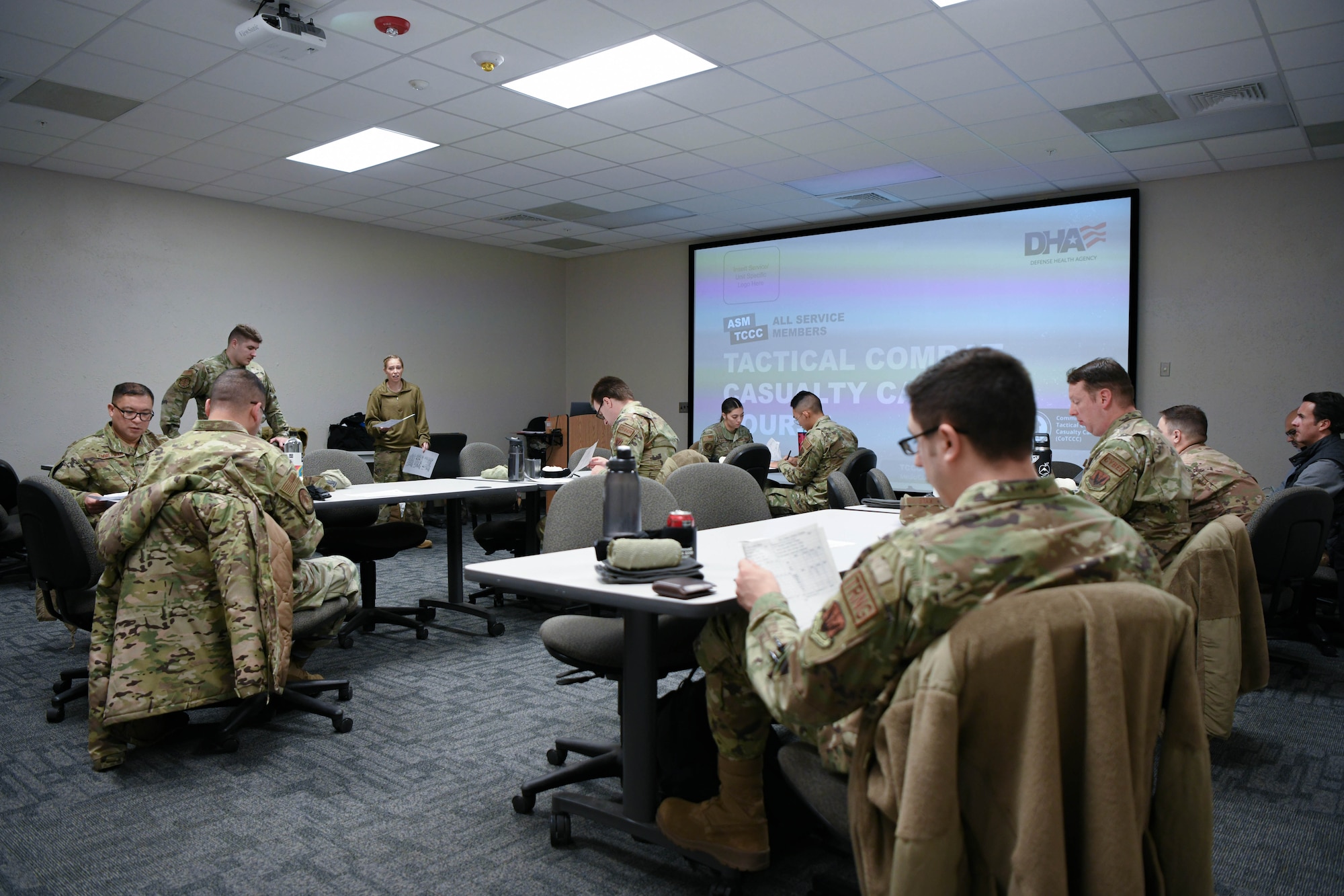 A group of people in military uniforms sit around tables in a classroom.