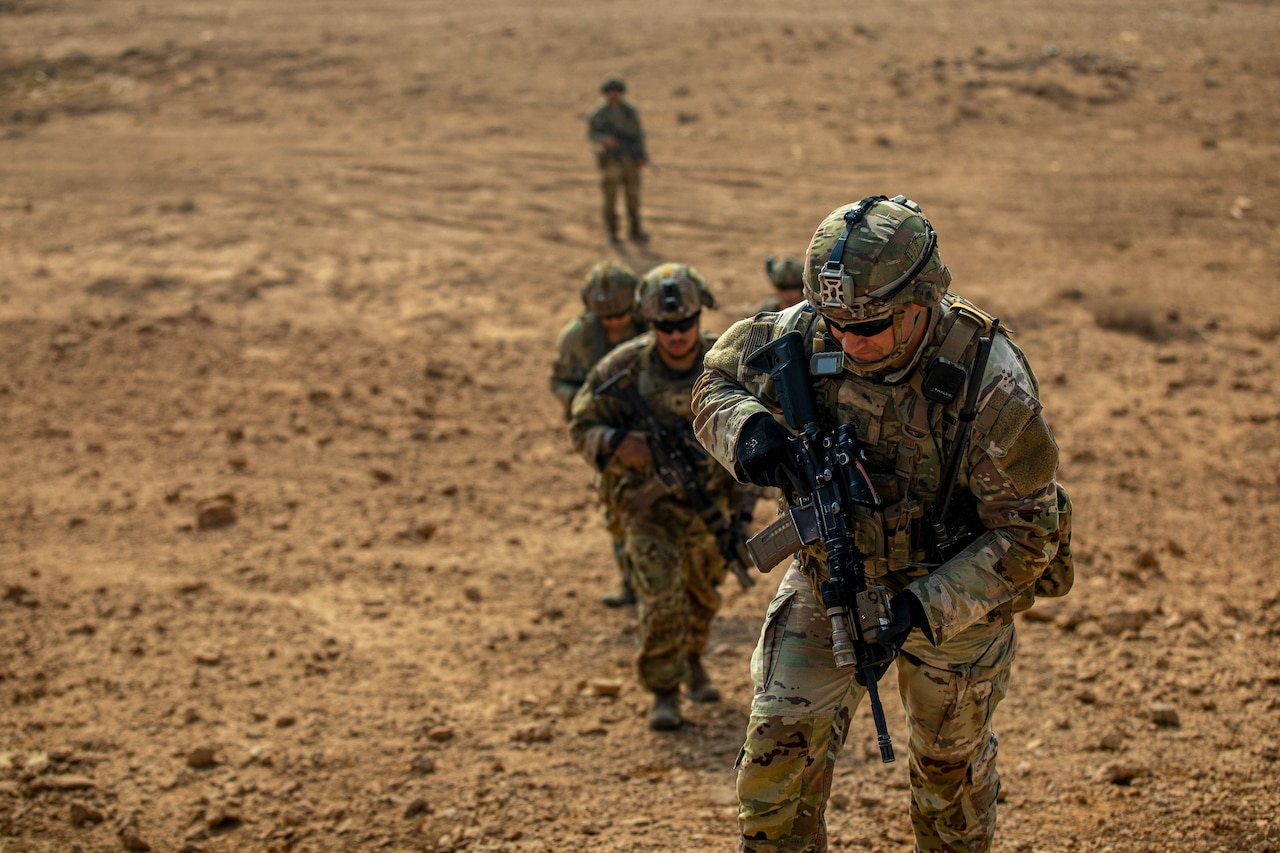 Soldiers carry rifles across a dirt field.
