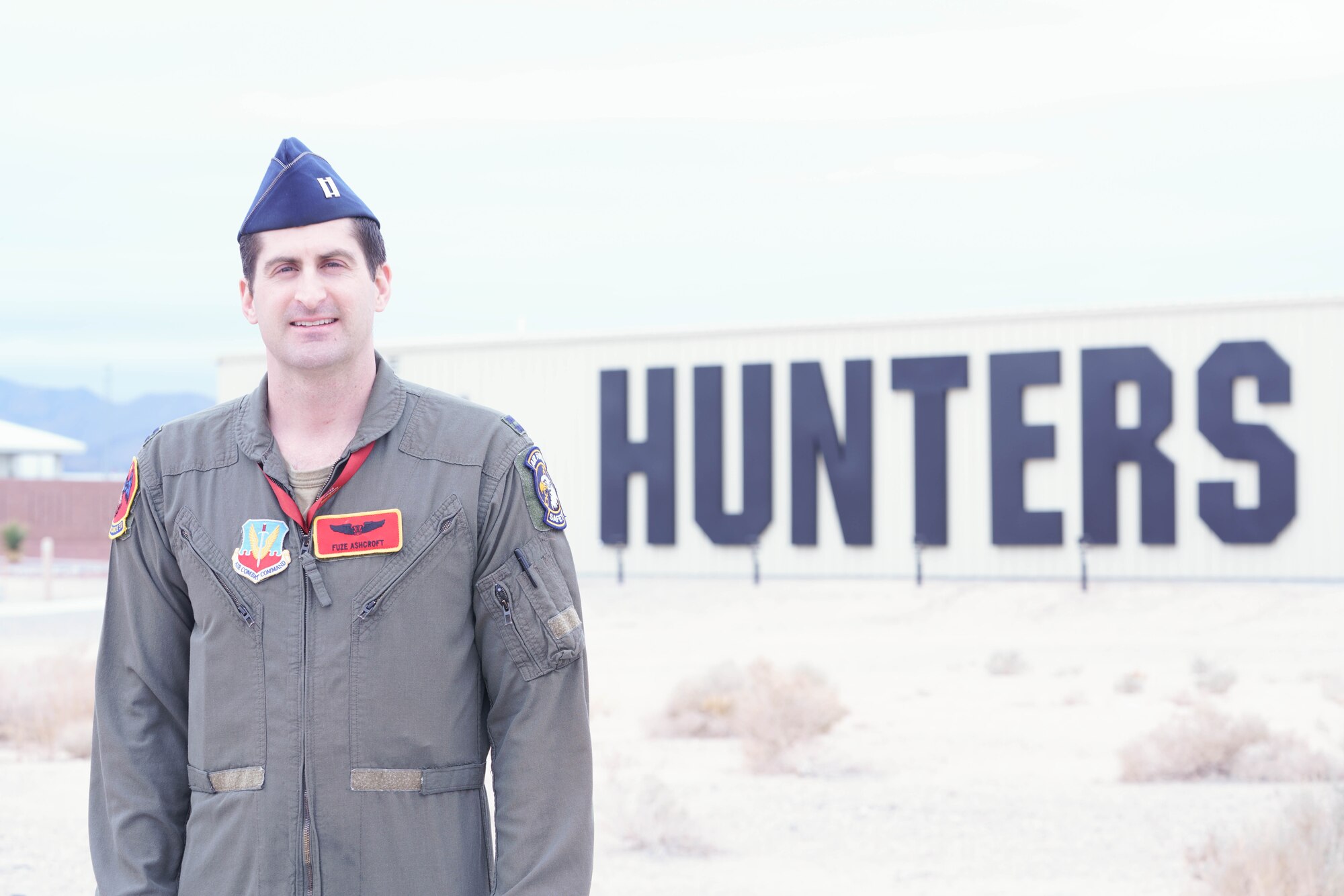 A man in a military flight suit stands in front of a building with the text "Hunters" on the side of it.