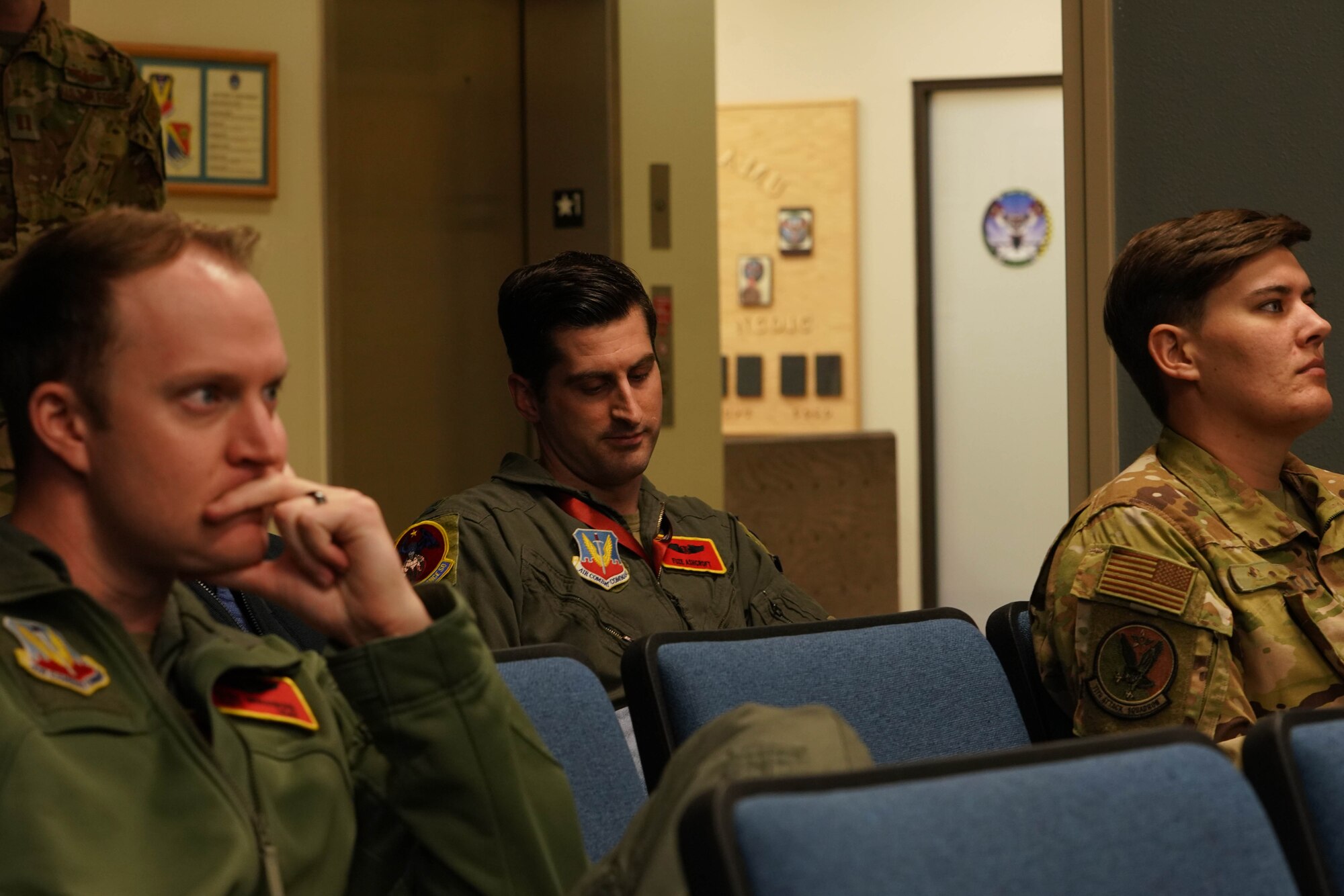 A man in a military flight suit takes notes sitting in an auditorium in between two other people in military uniforms.
