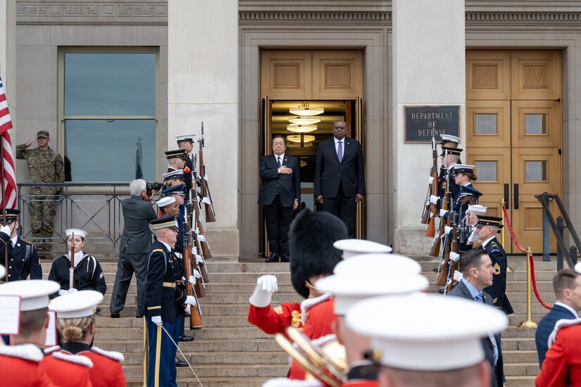 A ceremonial guard stands at attention while two men in the background stand and look on.  A man in the right foreground renders a salute.