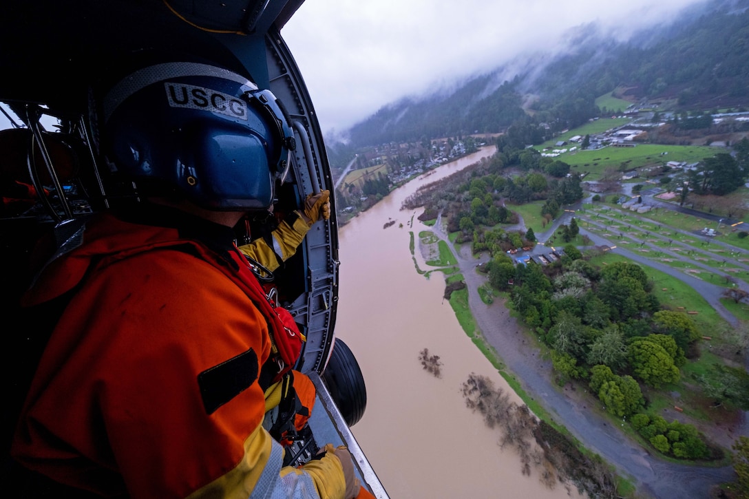 A coast guardsman looks out from a helicopter flying over a flooded region.