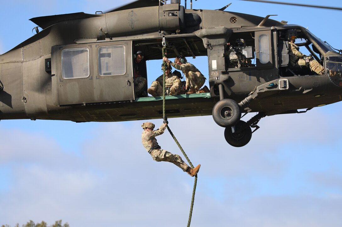 A uniformed service member descends from a helicopter using a rope as others look on.