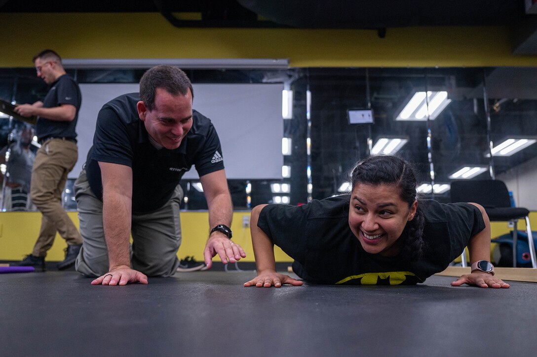 A service member smiles as she performs pushups in a gym.