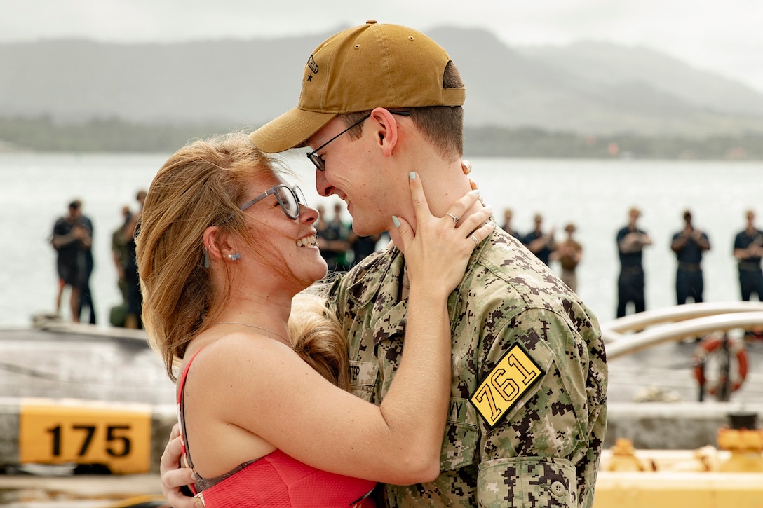 A sailor smiles at his wife as fellow sailors stand in the background near a body of water.