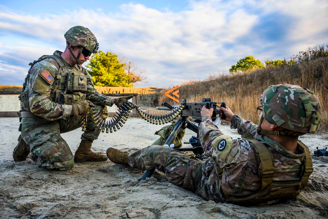 A guardsman lies on the ground as he fires a machine gun; another guardsman loads bullets into the weapon.