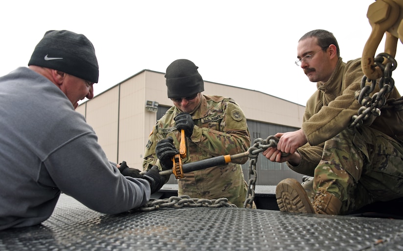 A U.S. Air Force airman, a U.S. Army soldier, and a civilian employee work together to detach a chain lock from a vehicle.