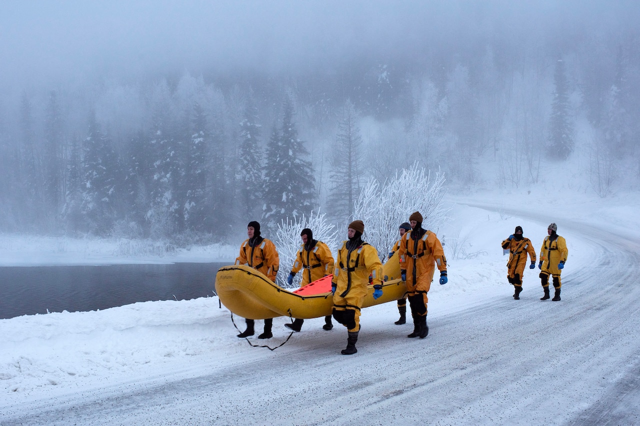 Firefighters in yellow dry suits carry an inflatable raft on a snow-covered road by a lake.