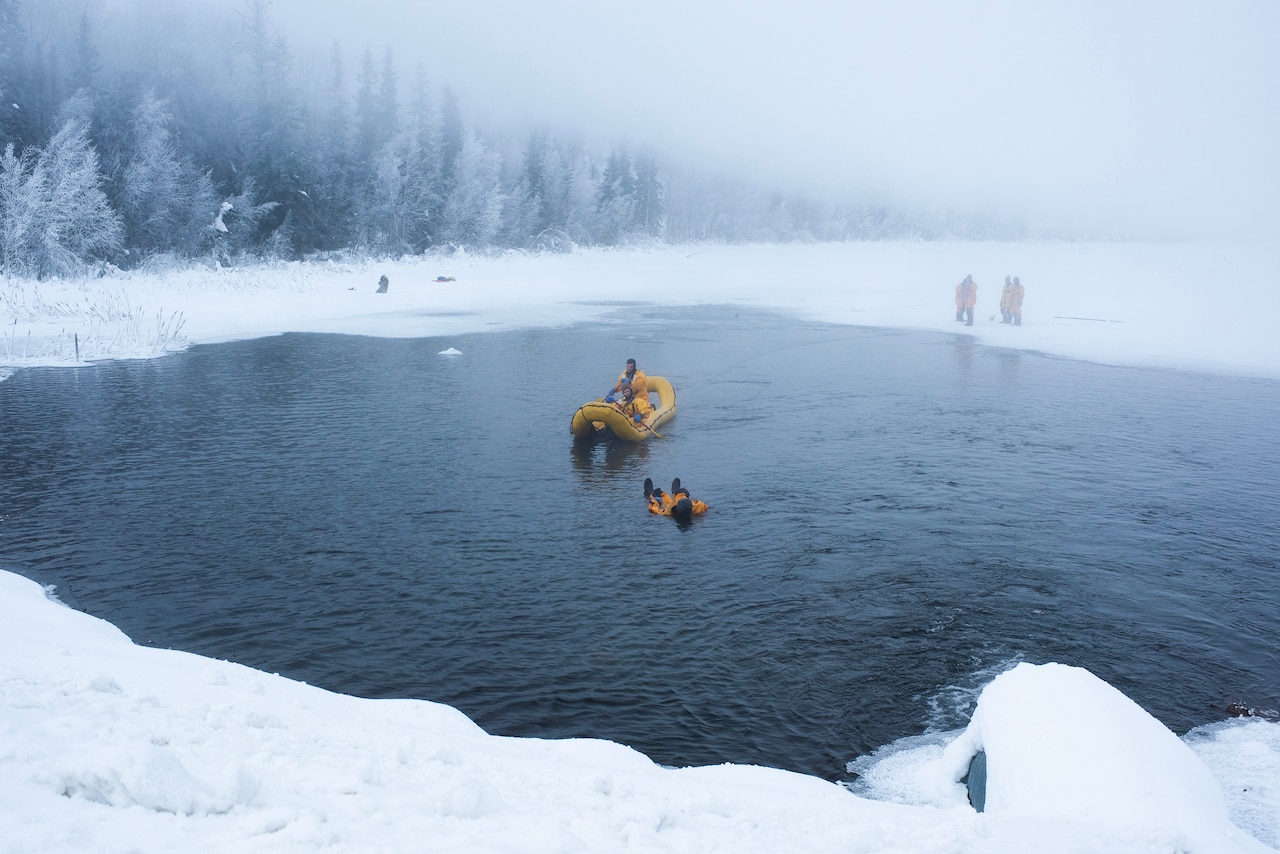 Firefighters in a yellow raft paddle toward another firefighter lying in a lake surrounded by snow.