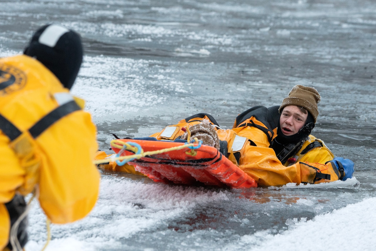 Two firefighters on a sled-type contraption gets pulled out of icy water by another firefighter.