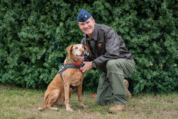 Man in flight suit crouches next to dog in front of tree