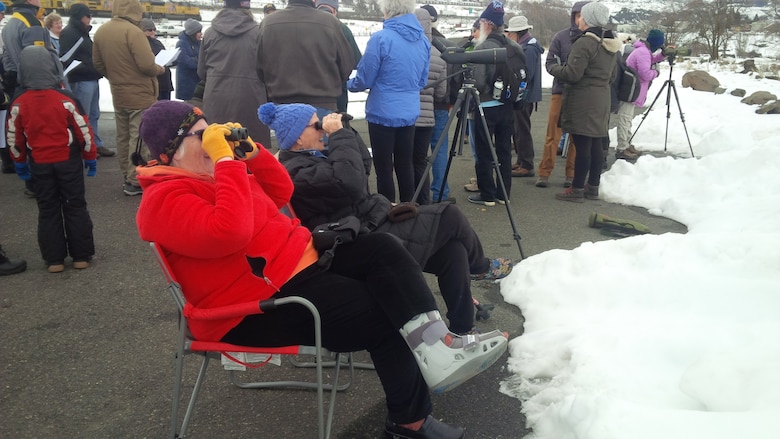 Two smiling people in winter coats and caps sit and look through binoculars on a winter day. Snow is on the ground just in front of the two people, and behind them a crowd of other people.