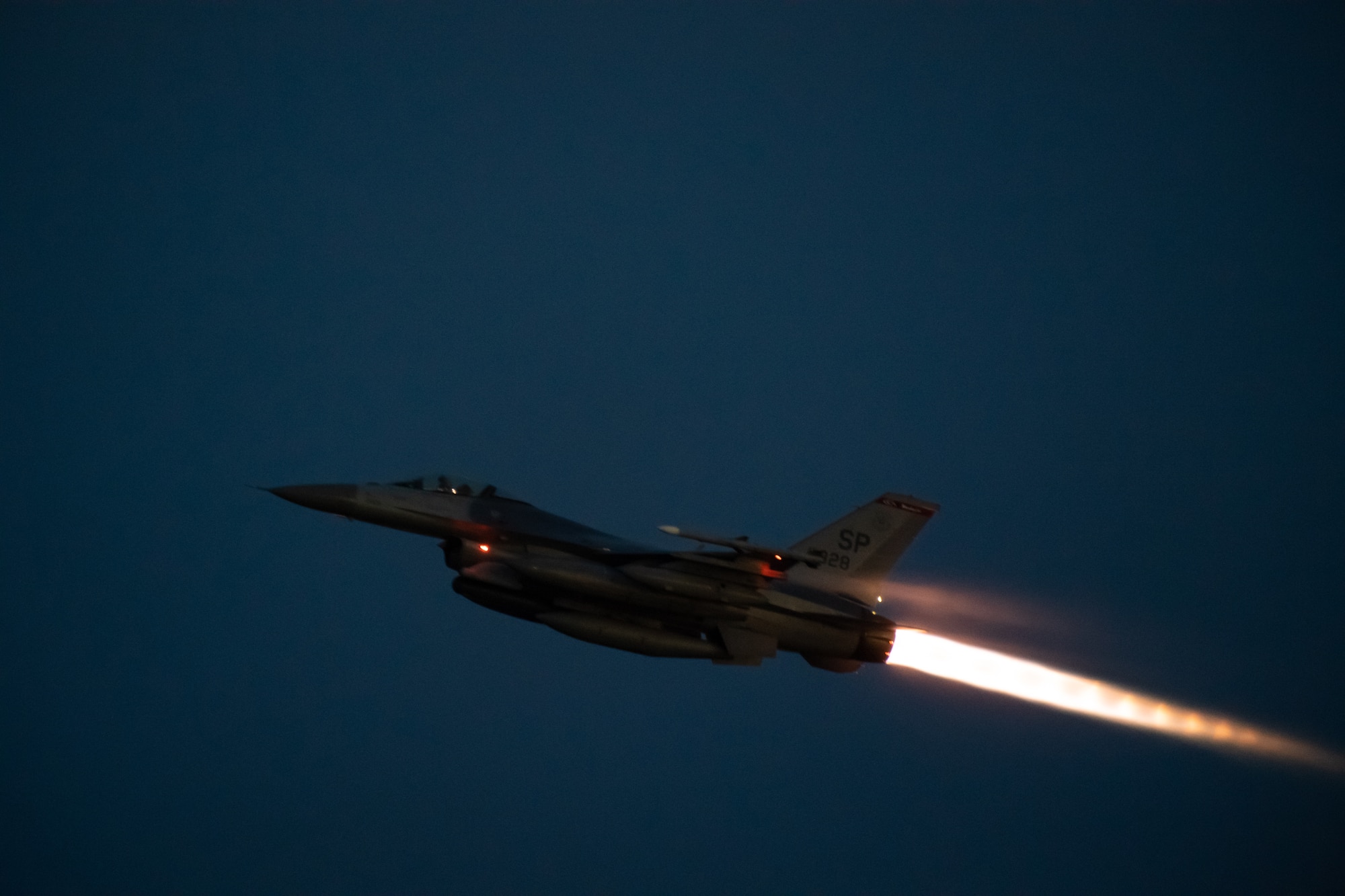 An F-16 flies across the night sky with its afterburner engaged.