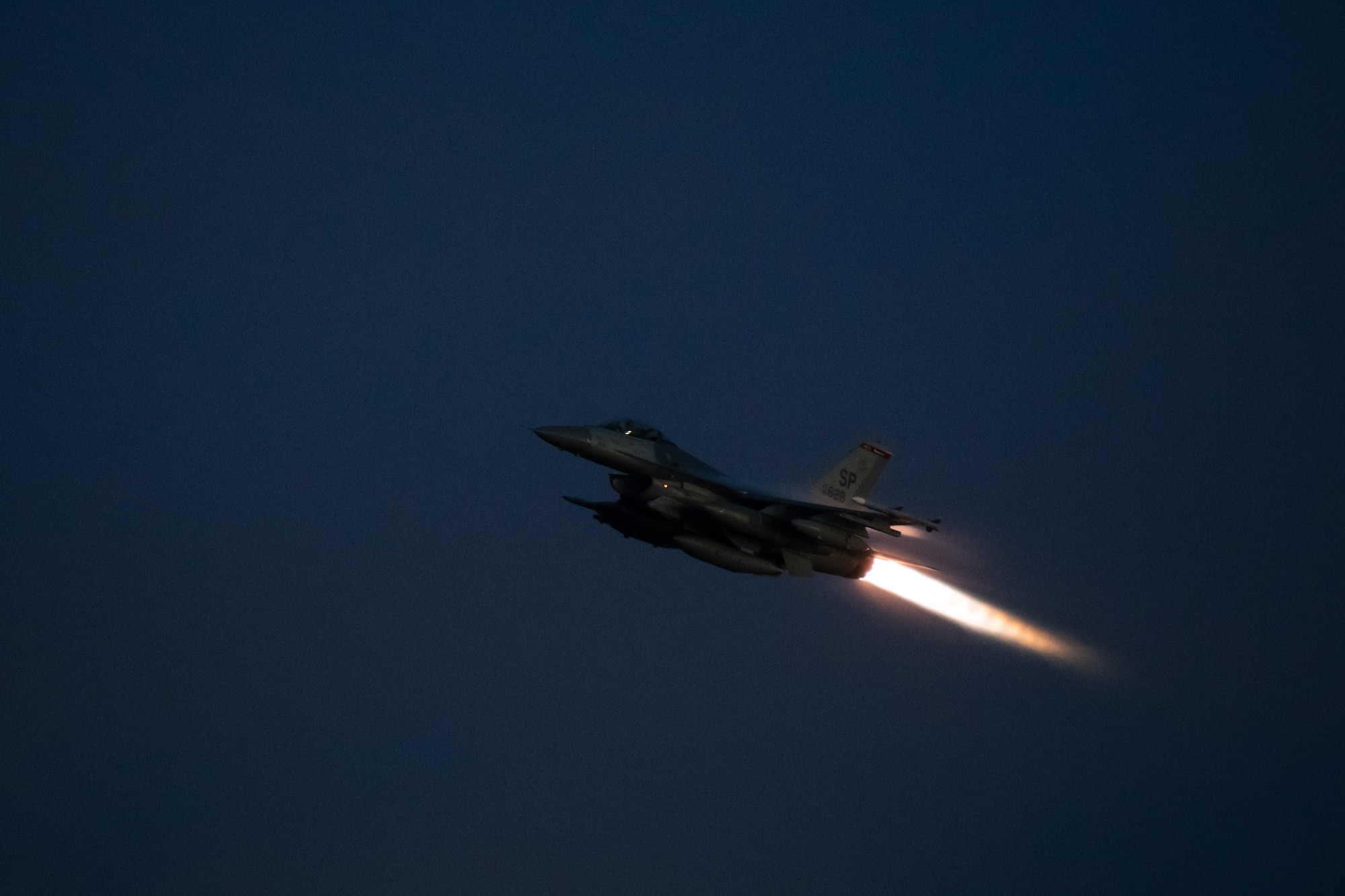 An F-16 flies across the night sky with its afterburner engaged.