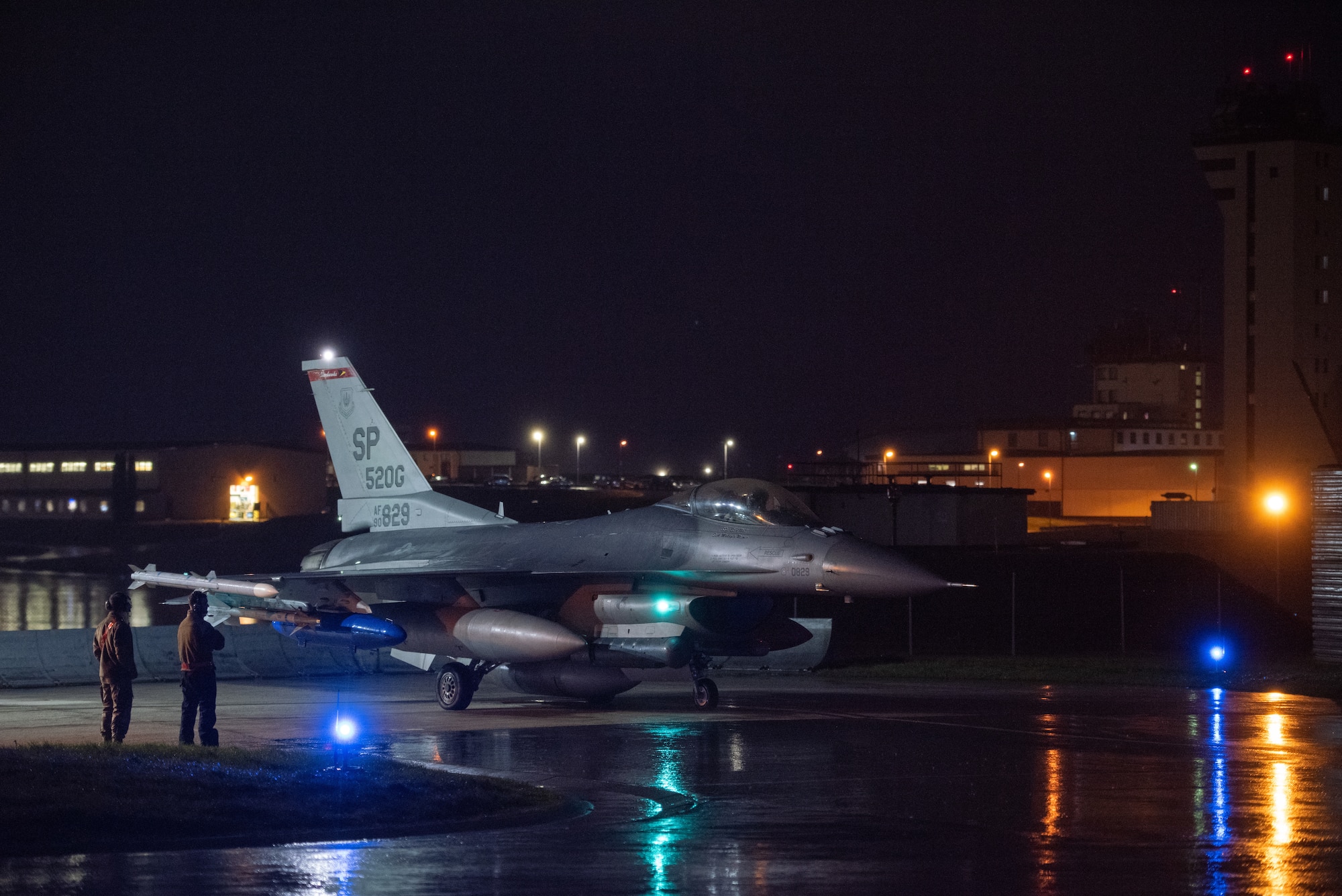 Lights reflect off of the wet flightline as an F-16 taxis across the flightline at night.