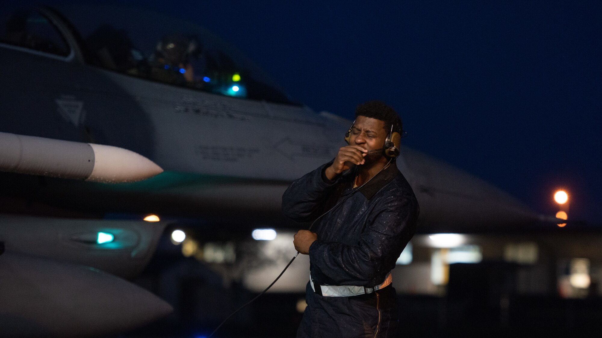 An Airman talks on a headset with an F-16 behind him in the background.