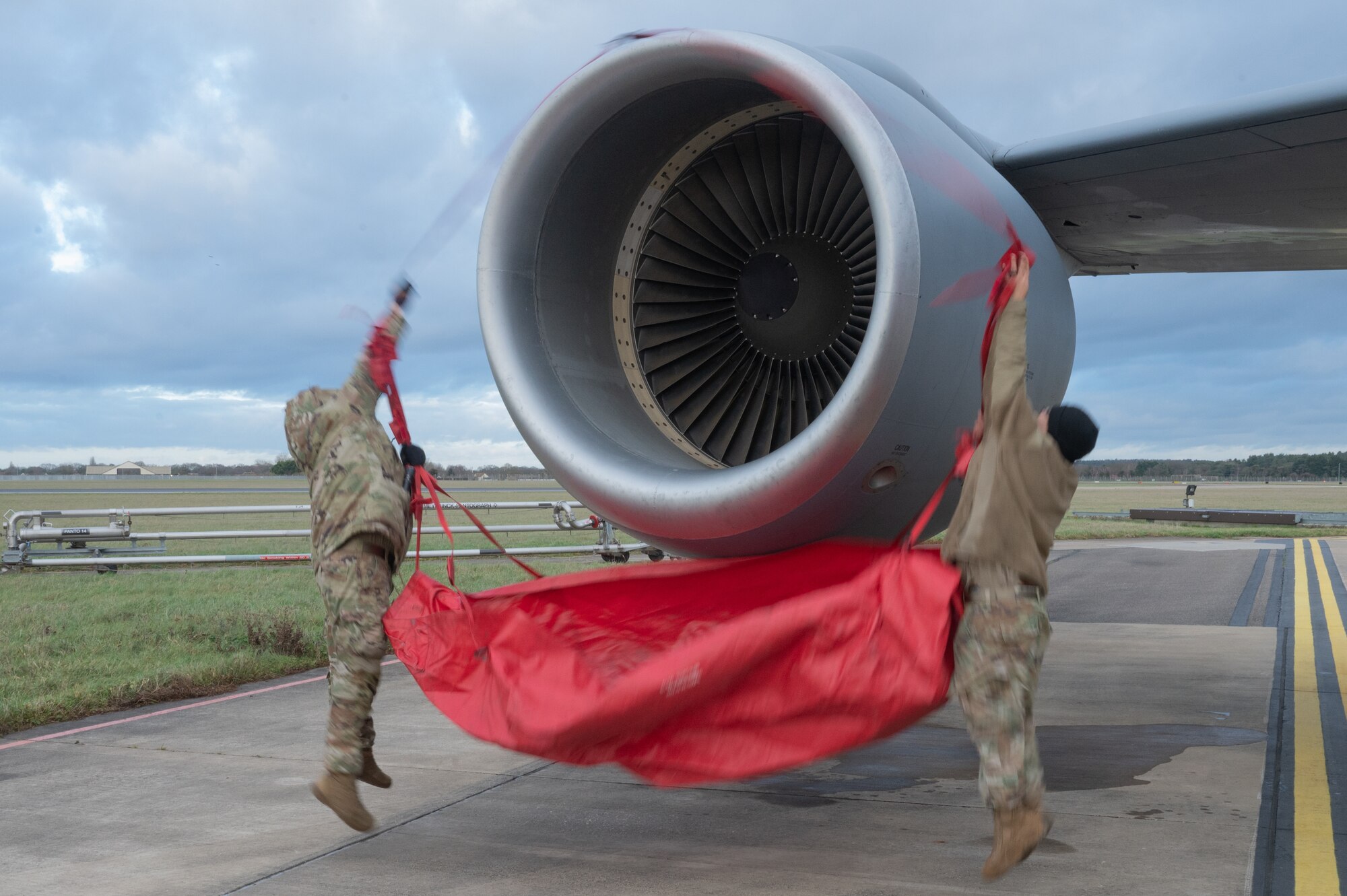 U.S. Air Force Senior Airman Eric Ridout, left, 100th Aircraft Maintenance Squadron aerospace propulsion journeyman, and Tech. Sgt. Angel Miranda Martinez, right, 100th AMXS aerospace propulsion NCO in charge, jump to get the top engine cover strap over the KC-135 Stratotanker aircraft engine on the flightline, Jan. 9, 2023, at Royal Air Force Mildenhall, England.