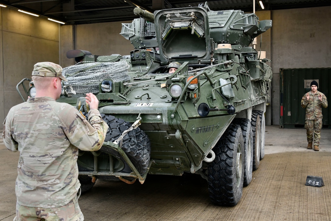 Two soldiers guide a military vehicle -- one standing in front of it and one behind it.