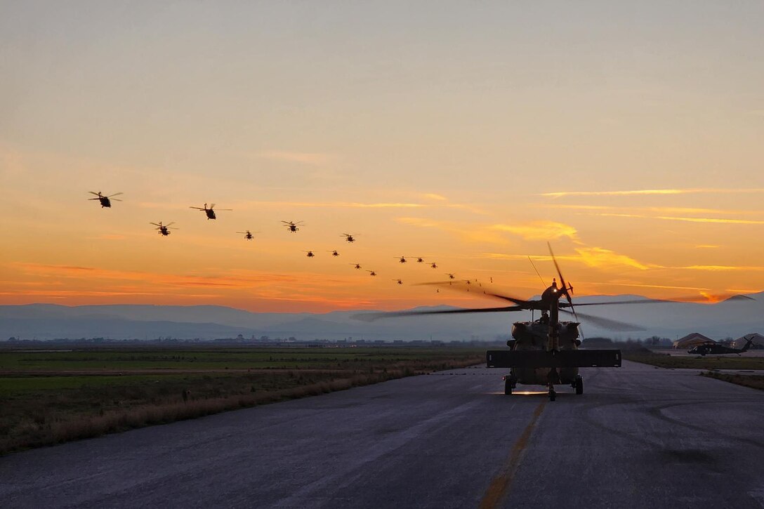 Helicopters fly in formation in a pinkish sky as one sits on a flight line.