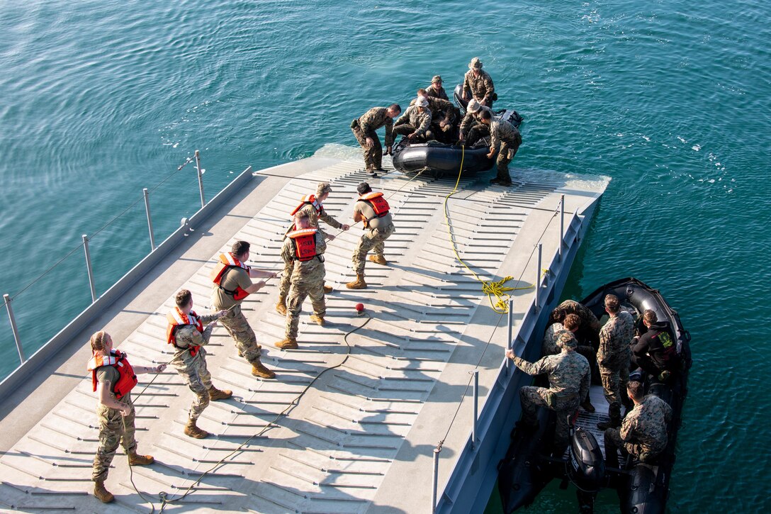 Marines pull a rope tied to a rubber craft as other Marines pull the craft onto a  ship's deck.