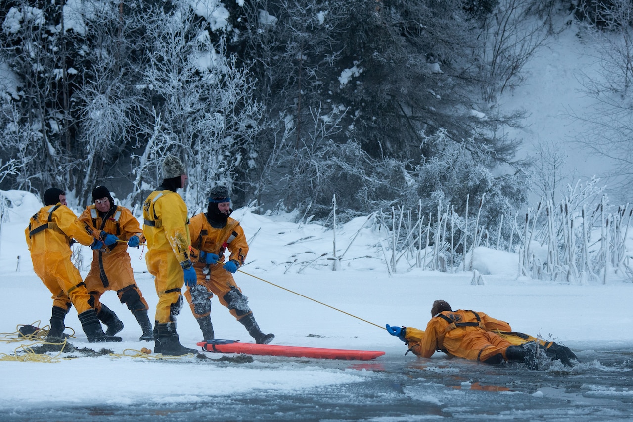 Four firefighters standing on snow pull a fifth out of an icy body of water using a line.