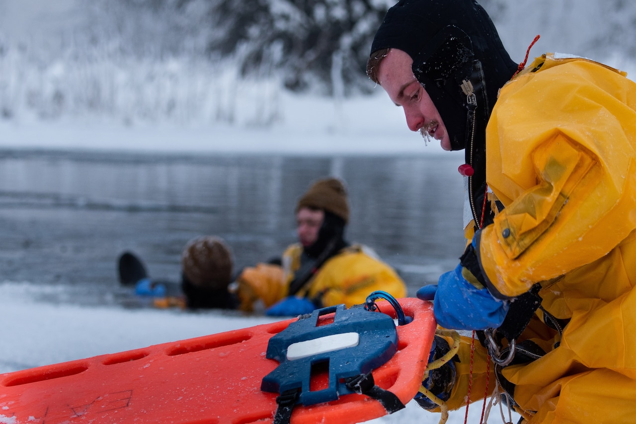 A firefighter in yellow dry suit handles a orange litter while others sit in icy lake.