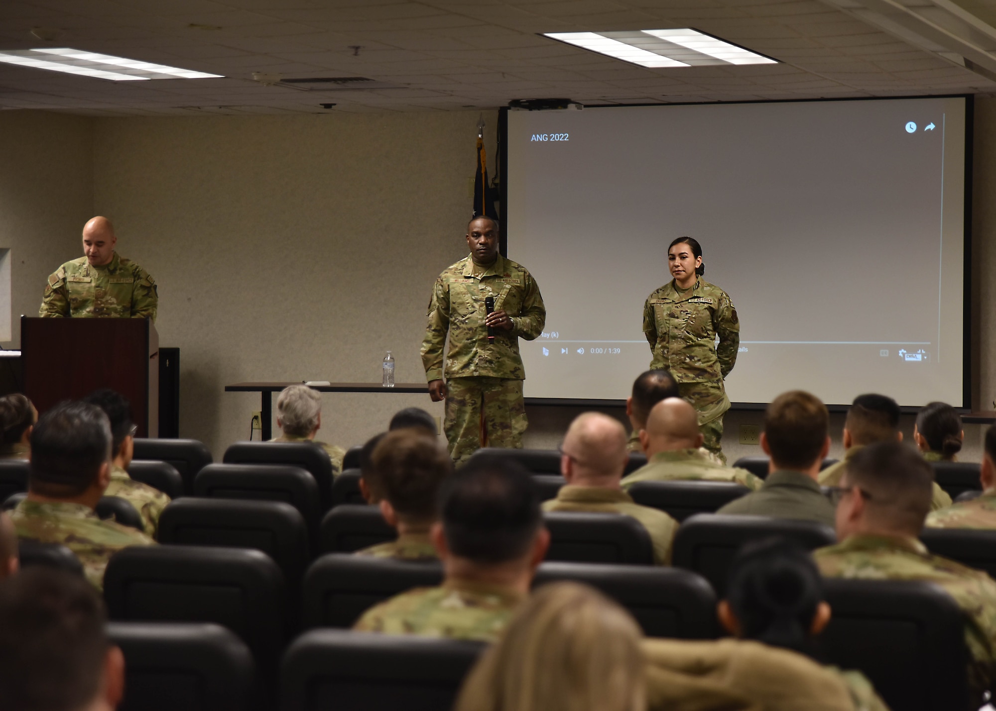 A man and woman wearing green camouflage uniforms stand in front of a room of people, who are also wearing green camouflage uniforms.