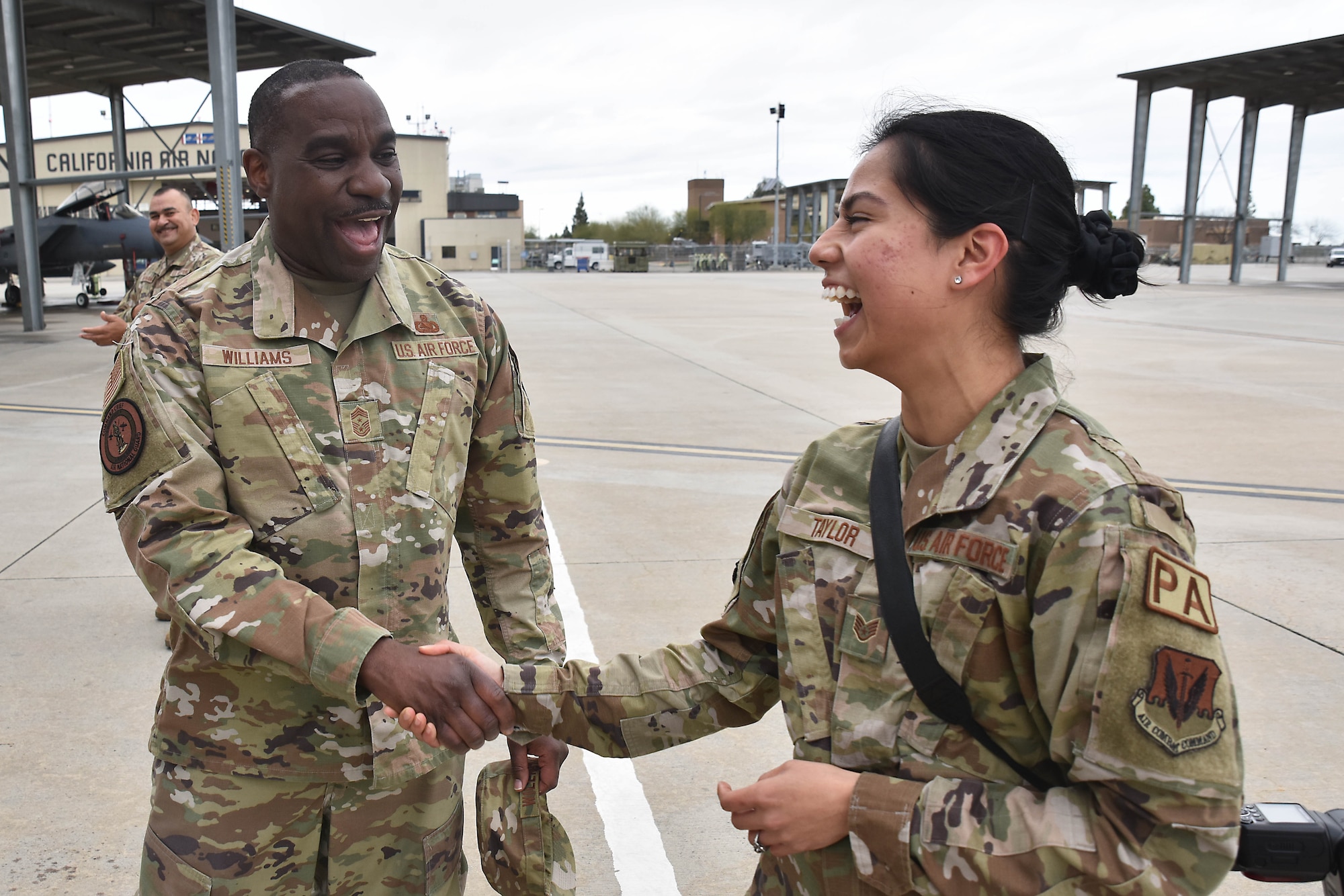 A man and a woman wearing green camouflage uniforms laugh while shaking hands