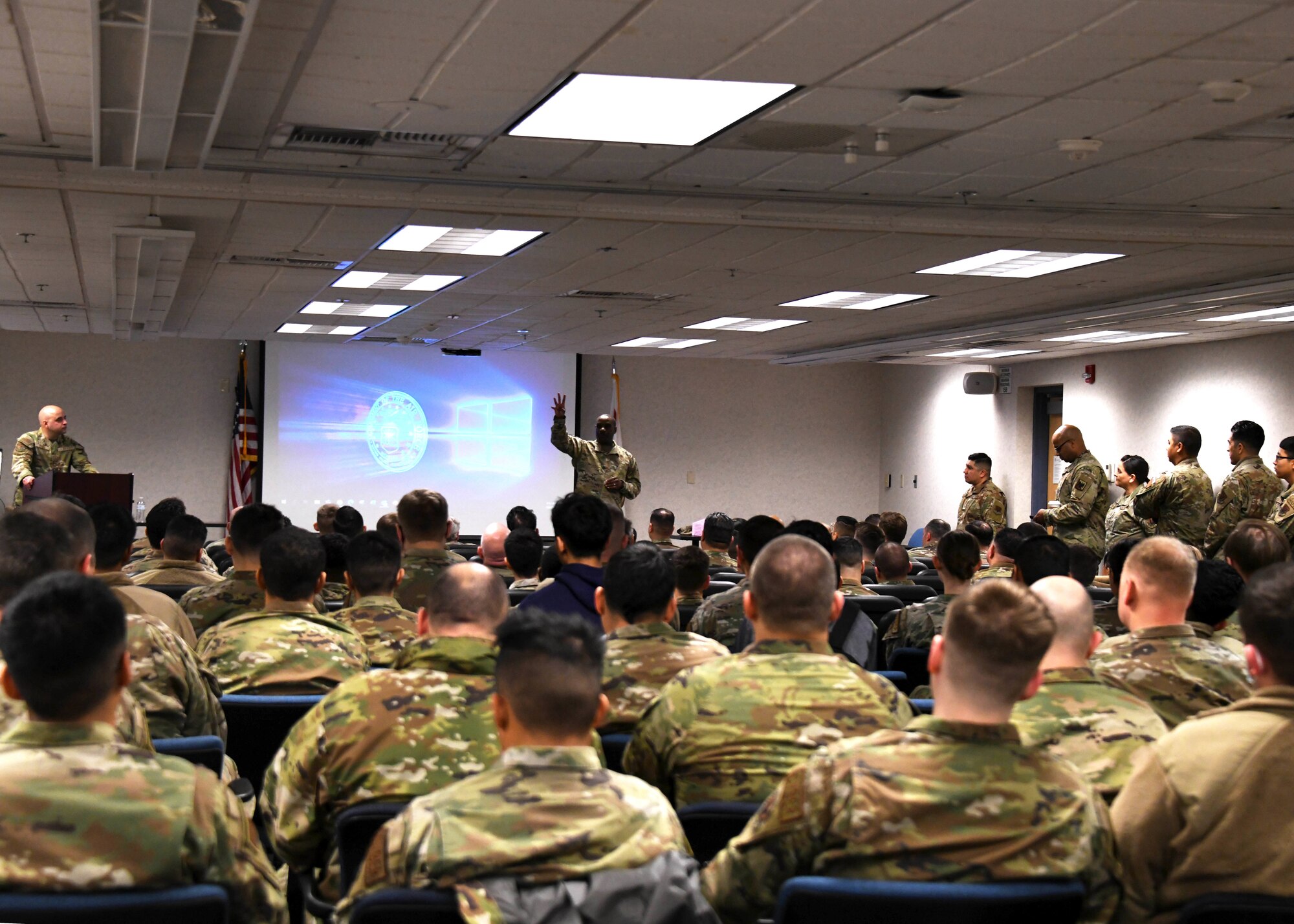 A group of people wearing green camouflage uniforms sit toward a man wearing a green camouflage uniform speaking to them