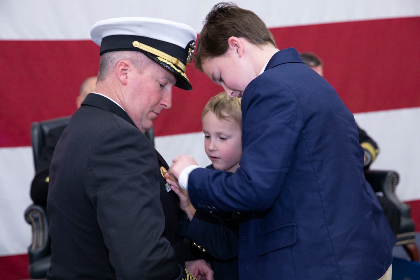 Capt. Dan Pugh, incoming Commander, Fleet Logistics Support Wing (CFLSW), kneels for his two sons to pin his command pin to his service dress blue jacket during the CFLSW change of command ceremony.
