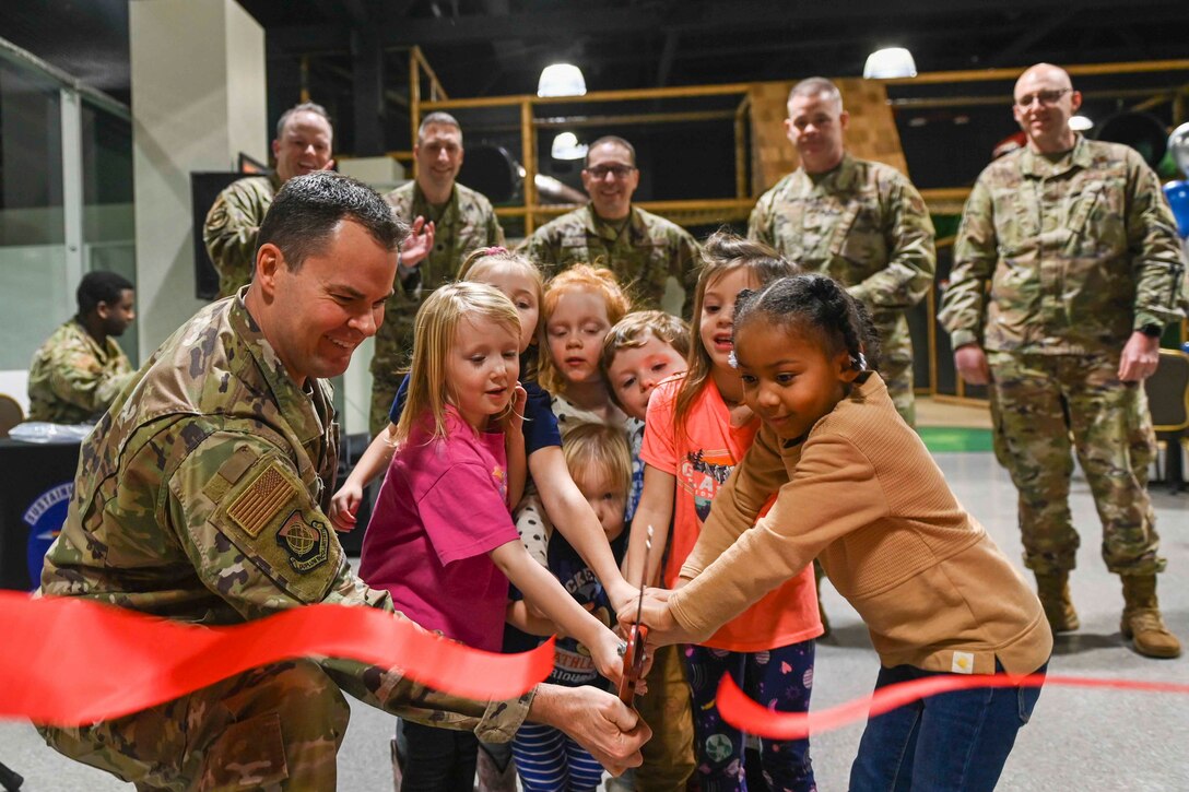 An airman helps a group of children cut a ribbon as other airmen watch.