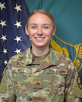 Women wearing U.S. Army uniform standing in front of two flags.