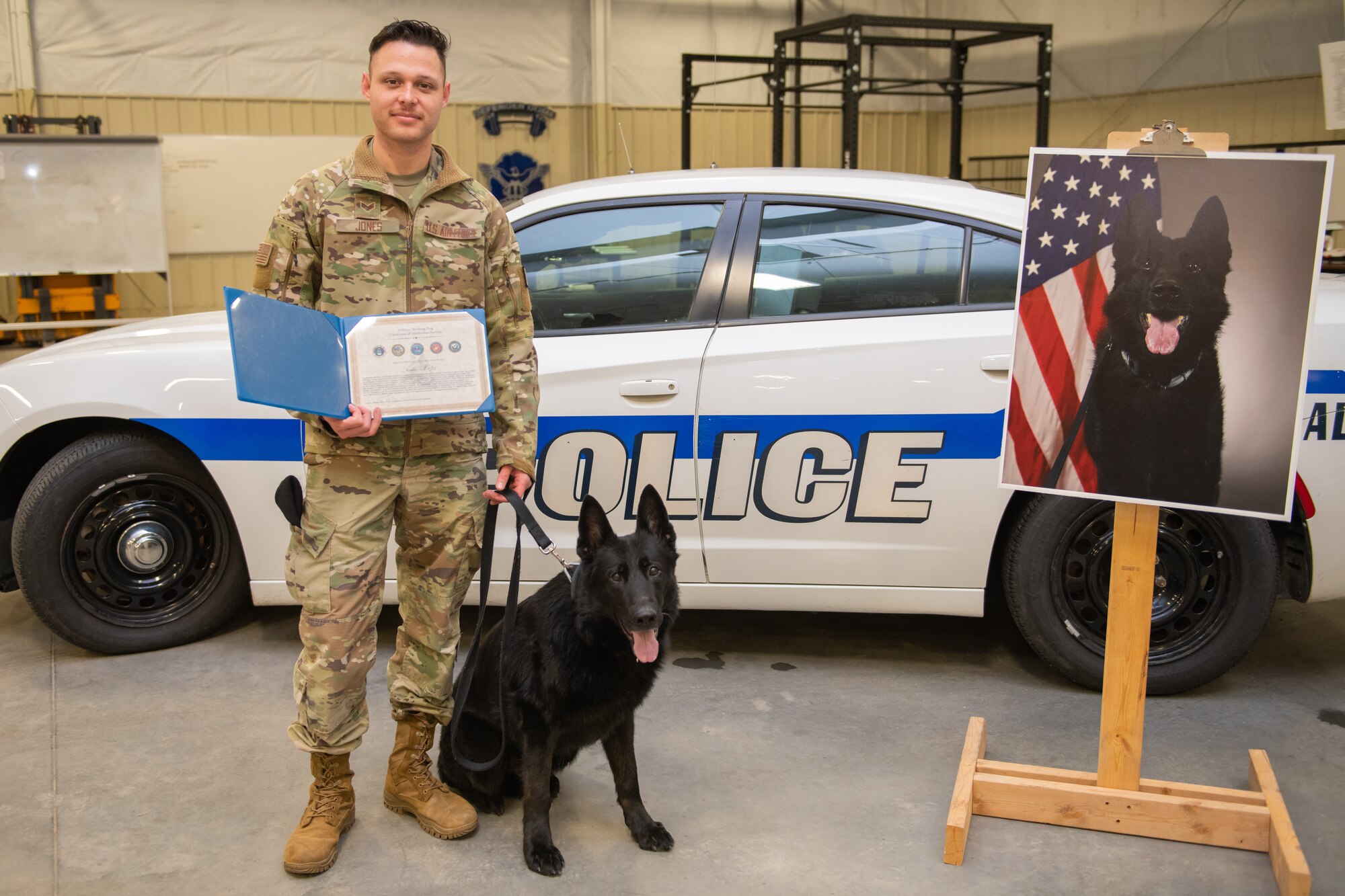 A man in uniform poses with a black dog in front of a police car, holding a certificate.