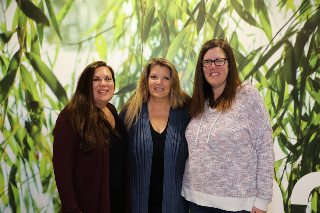 Three women pose for a photo in front of a green and white background.