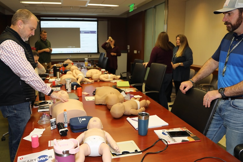 Men and women stand around a table practicing CPR and first-aid training in a dimly lit room.