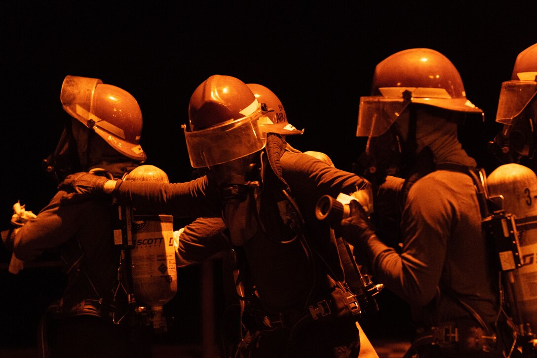 Sailors in firefighting gear stand in a line in a dark space.