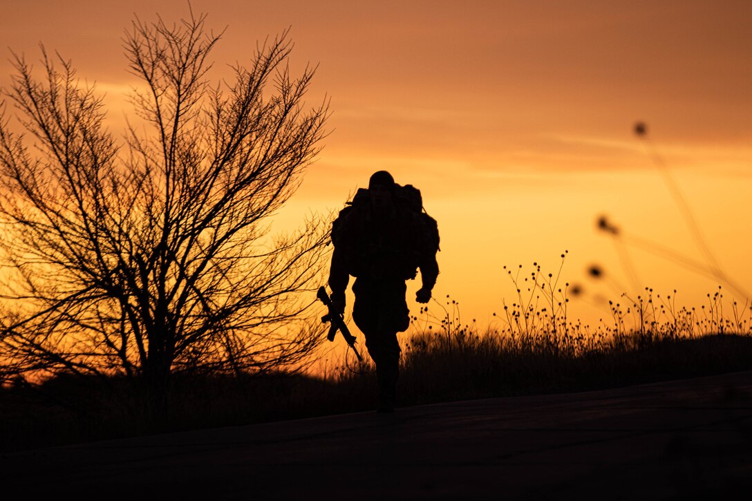 A soldier is shown hiking. The image is silhouette, illuminated by an orange sky.