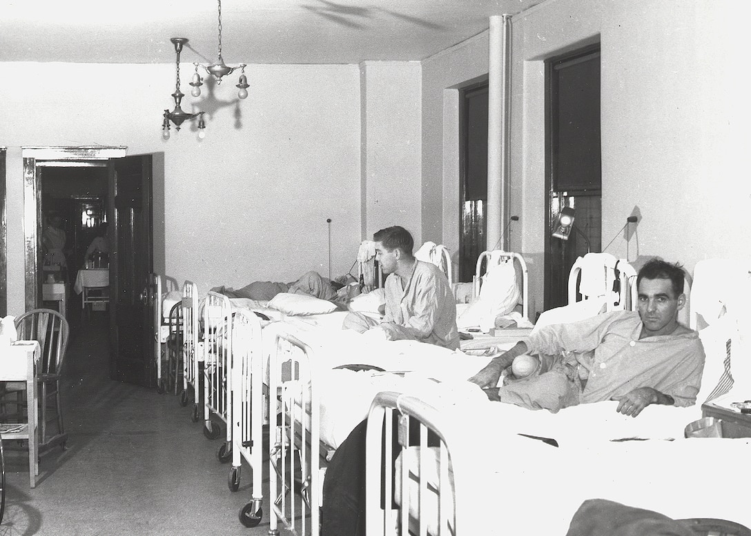 Black and white photo of patients in beds next to each other recovering from wounds. The Soldier nearest the camera is looking in to the camera, he is missing his lower right leg.