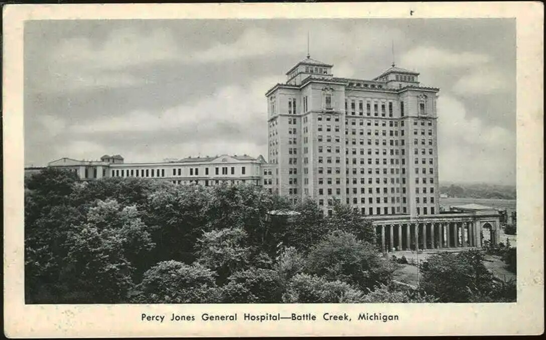 A WWII era postcard shows the Percy Jones Army Hospital building. The image is in black and white. in the foreground are trees that hide part of the six story building but the tower addition stands tall with 15 floors.