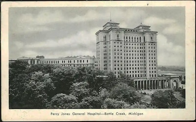 A WWII era postcard shows the Percy Jones Army Hospital building. The image is in black and white. in the foreground are trees that hide part of the six story building but the tower addition stands tall with 15 floors.