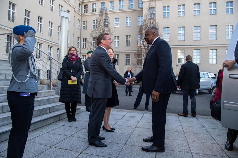 Two men shake hands in front of some steps as others stand around them.