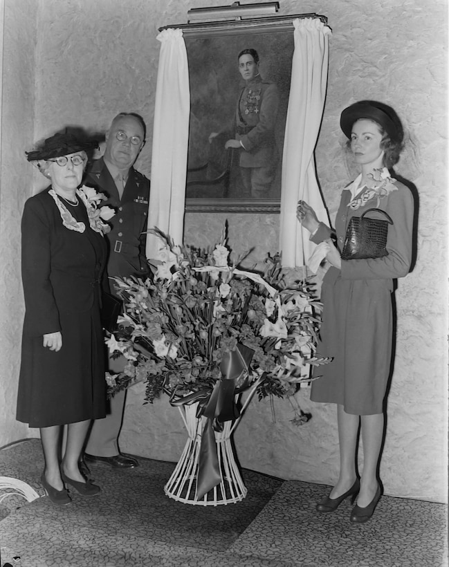 In a black and white photo three people stand next to a painted portrait of a man (Col. Percy L. Jones) in an old style Army uniform. two of the people are women, the widow of Percy Jones and their adult daughter (who is the tallest person in the photo) and an Army General stands next to Percy Jones' widow.