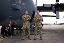 Two men in Air Force uniform stand in front of a large aircraft on concrete with more planes visible in the background.